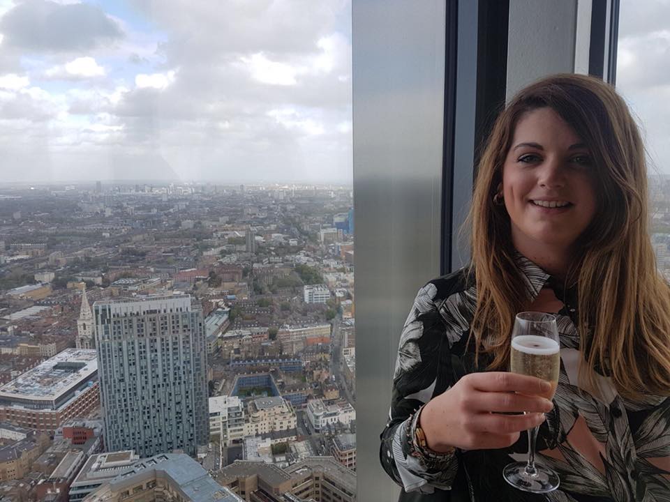 Linzi, a women with short brown hair, smiles at the camera, she wears a black blouse and holds a glass of champagne, she stands against a window that outlooks city buildings.