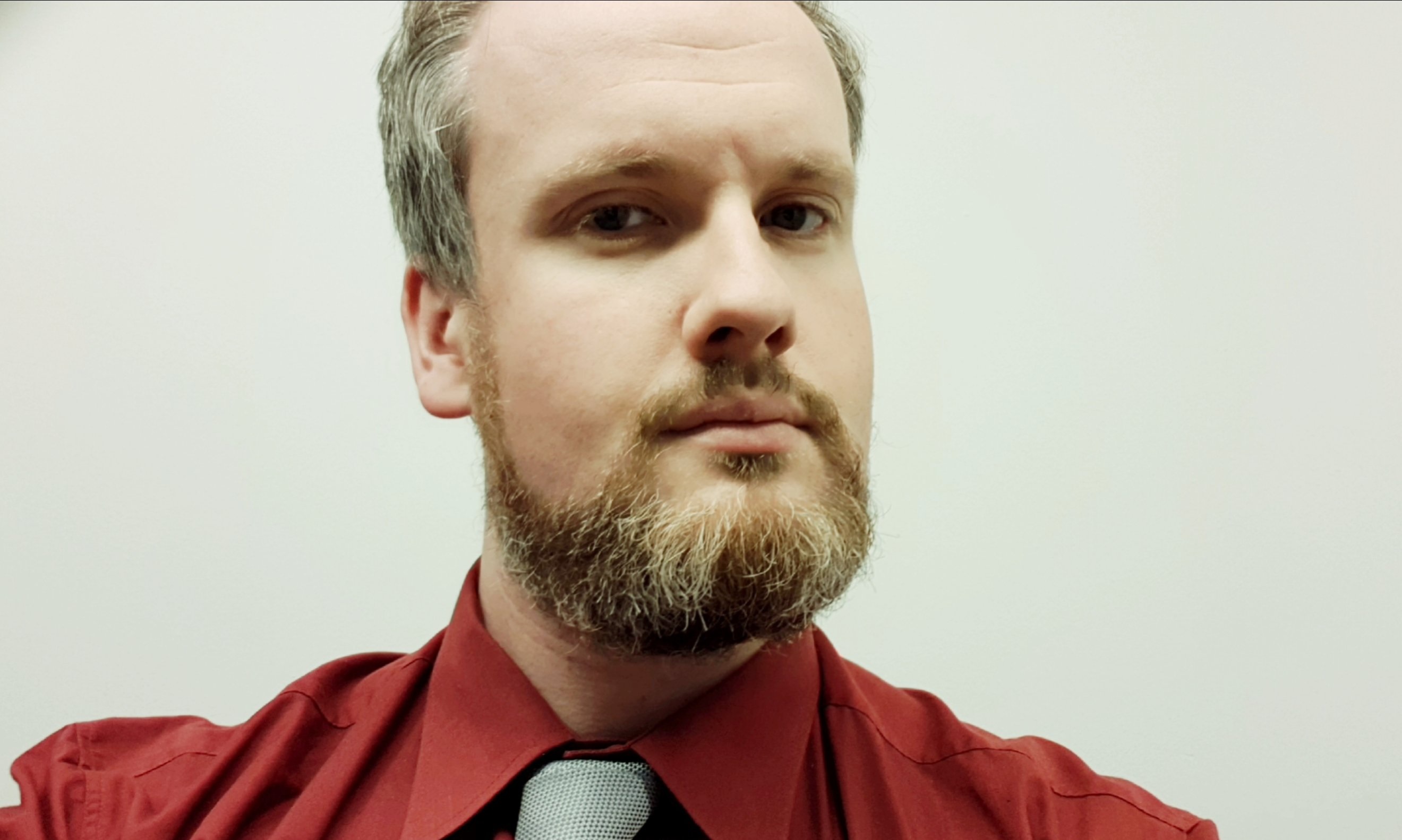 A portrait of Patrick who wears a red shirt and grey tie, he stands against a white background. 