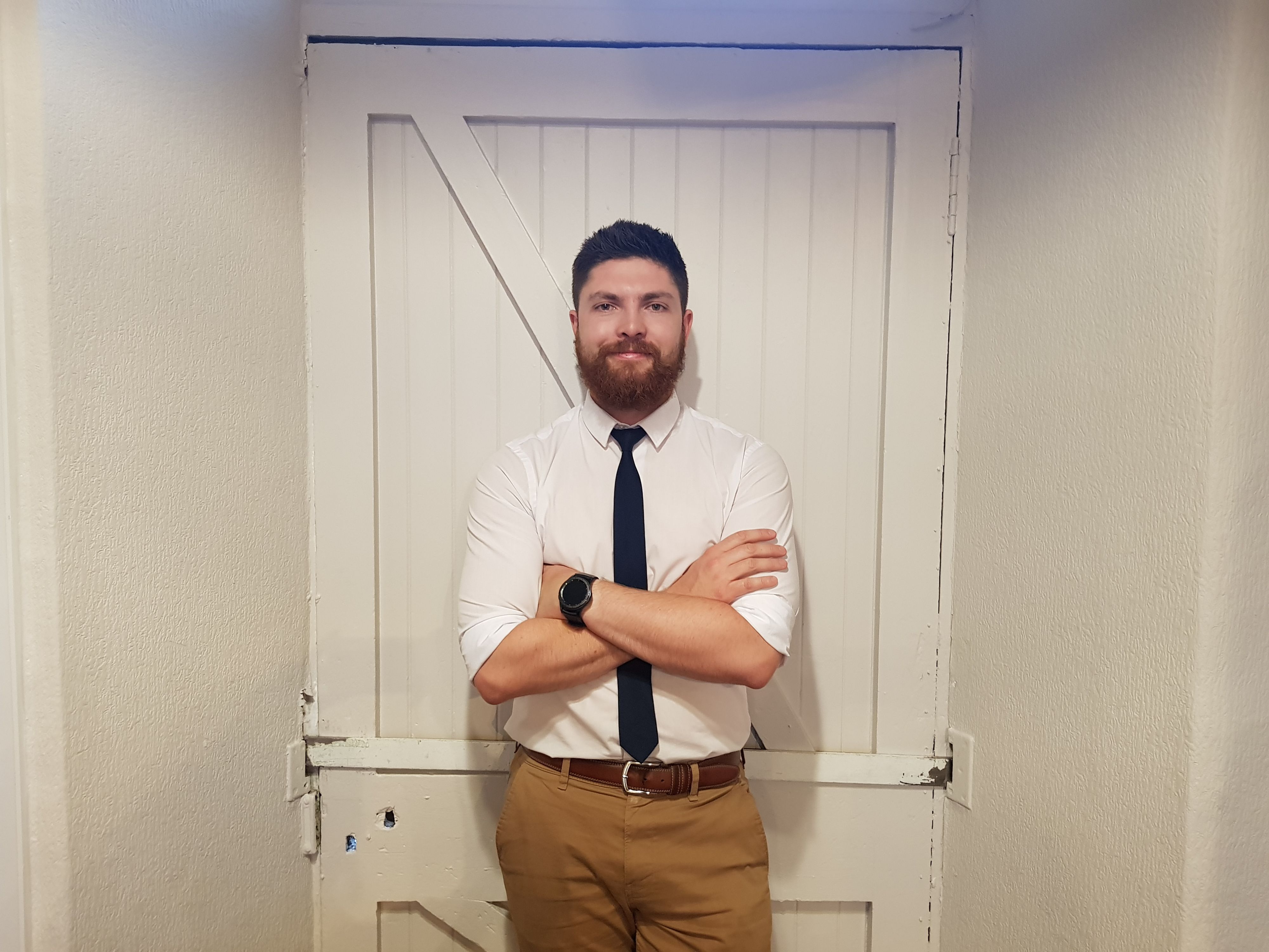 Man with a beard wearing a white shirt and black tie, standing with arms folded in front of a white wooden door.
