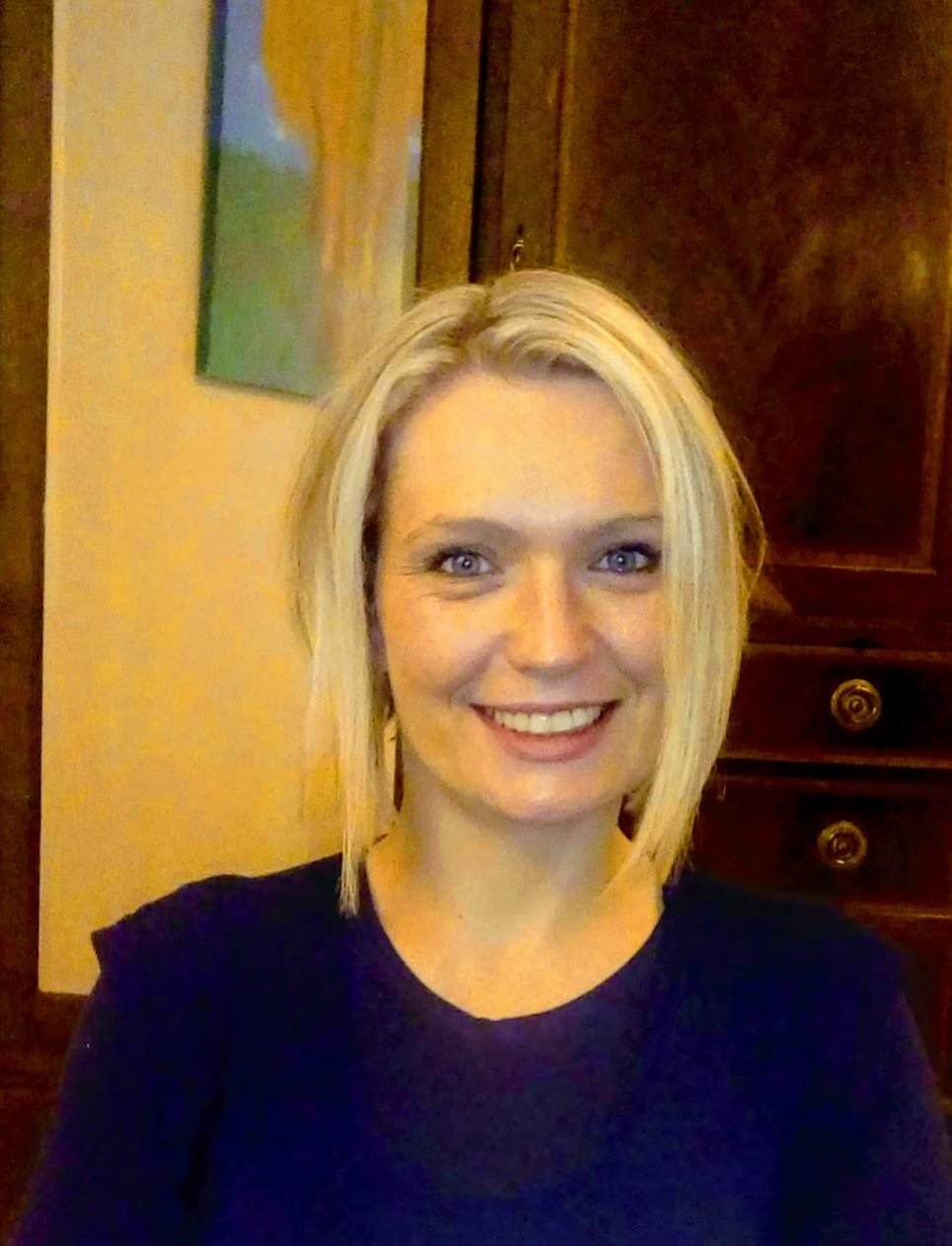 A woman with short blonde hair wearing a a blue top smiles at the camera, she poses against a brown wardrobe with cream walls in the background.