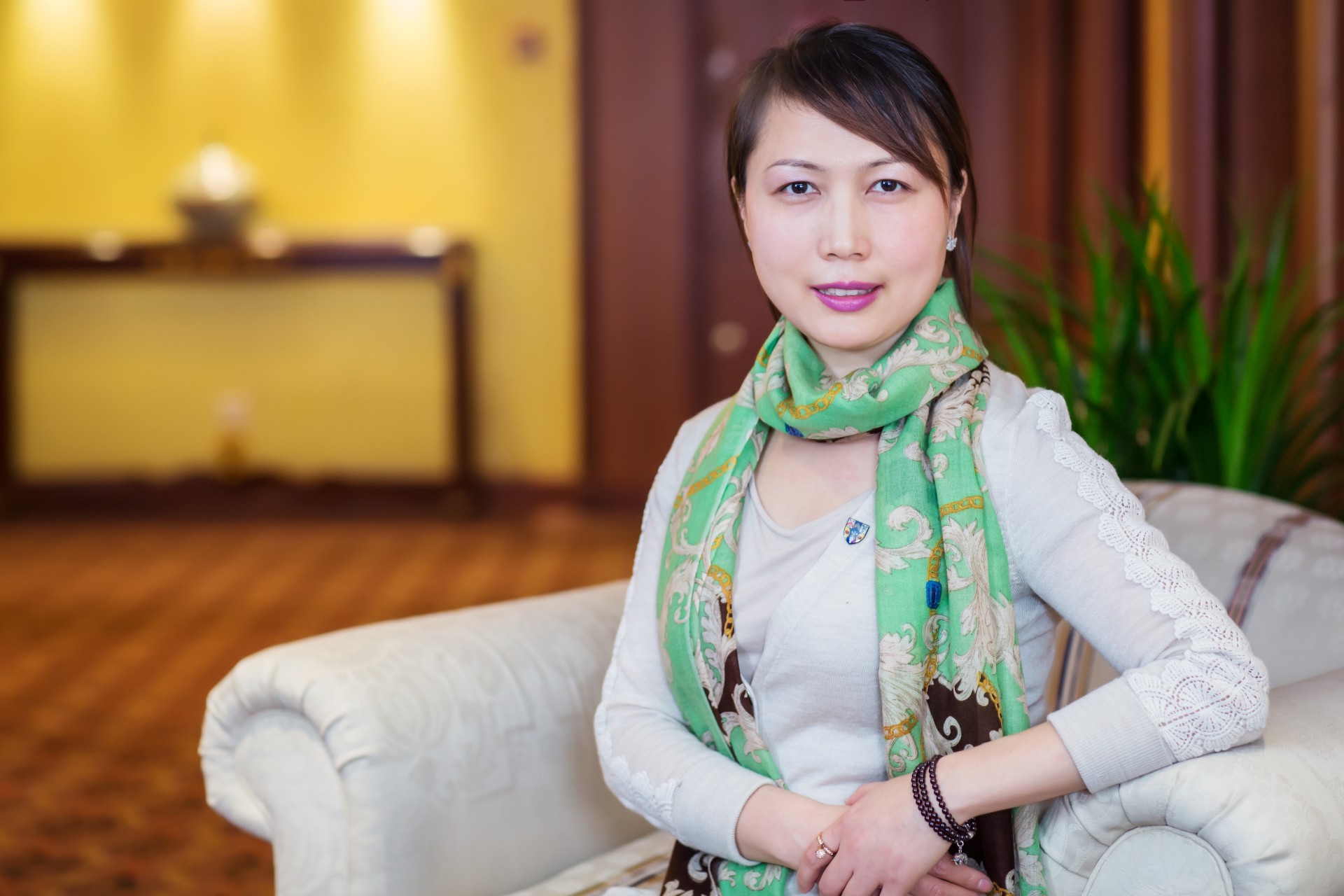A woman with black hair wearing a white jumper and green floral scarf smiles at the camera, she sits on a white chair. 