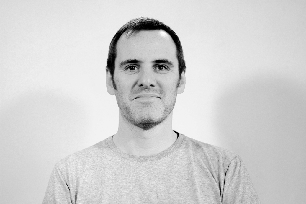 A back and white portrait of a man looking at the camera, he has dark hair and has a slight smile. 