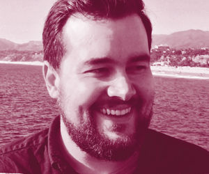 A close up headshot of a man with dark hair and a beard, he smiles off to the right of the camera, the background has ocean water and a beach. The image has a pink tint filter on it. 
