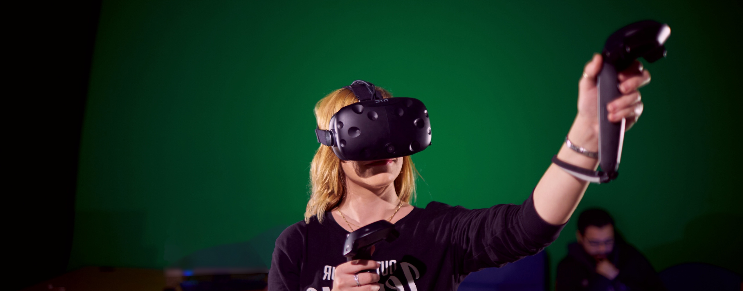 Student wearing VR headset and motion controllers engages with immersive virtual reality technology against a green screen backdrop, demonstrating interactive gaming or research applications.