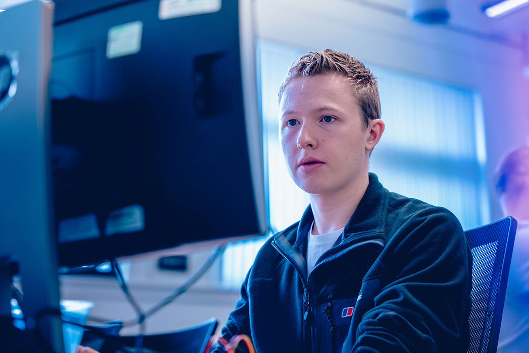 male with blonde hair wearing sitting behind a computer screen with a concentrated expression