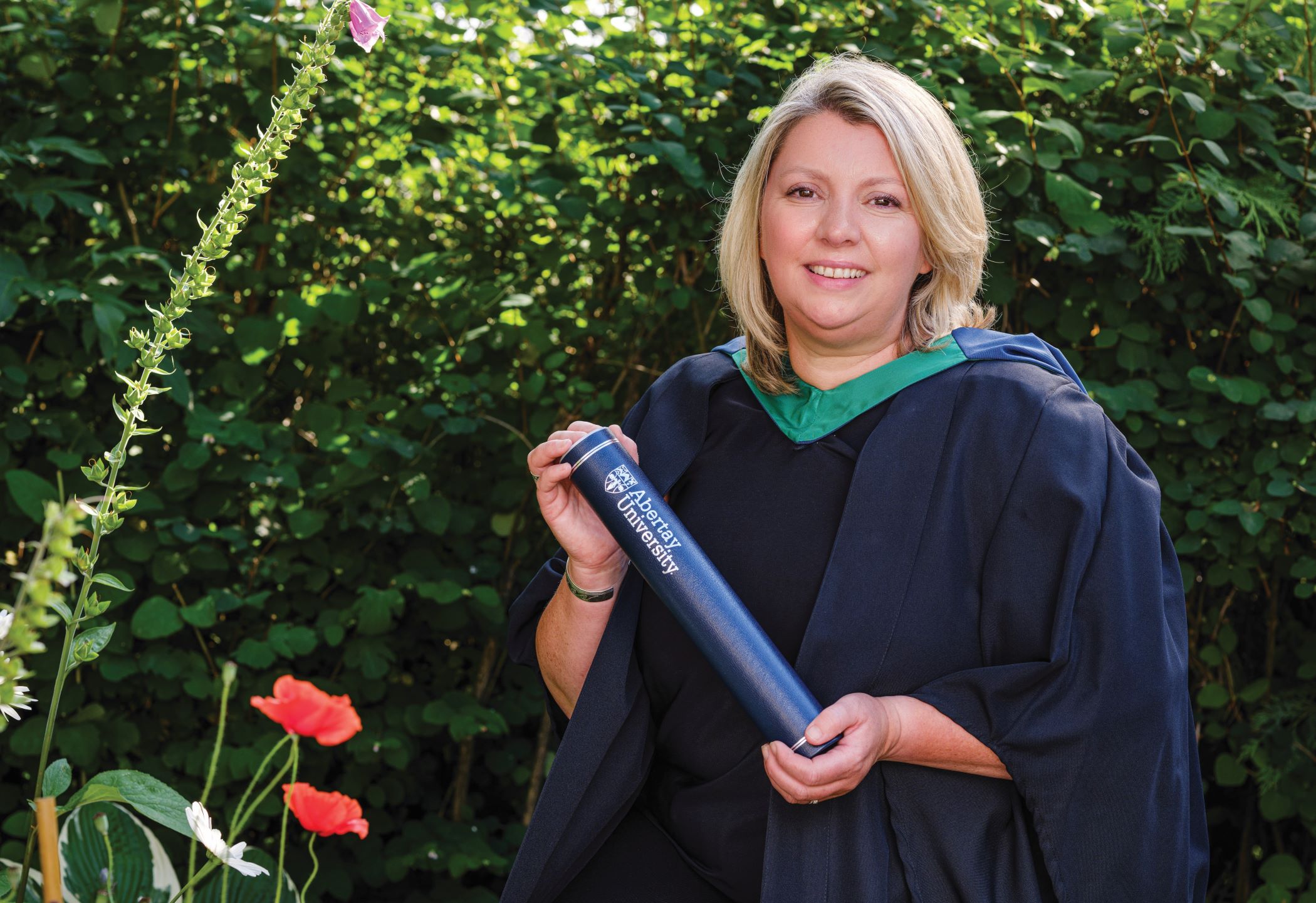 Kirstin is smiling with shoulder-length blonde hair wearing a dark graduation gown with a green hood, holding a dark blue rolled Abertay certificate case, standing in front of leafy green bushes.