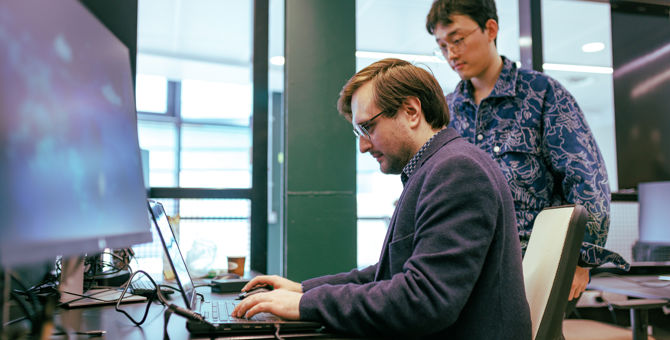 Two males working at a computer: one seated typing on a laptop while another stands behind looking at the screen