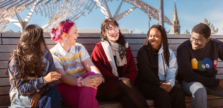 A group of students sitting on a bench outside smiling, behind them is blue sky and visible a metal whale sculpture at Dundee's waterfront.