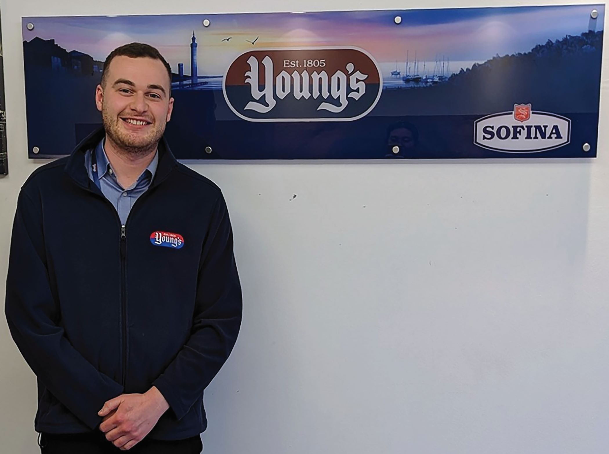 Malcom smiles at the camera against a white background with a sign that says 'Youngs'. He has dark hair and wears a navy blue 'Youngs' fleece.