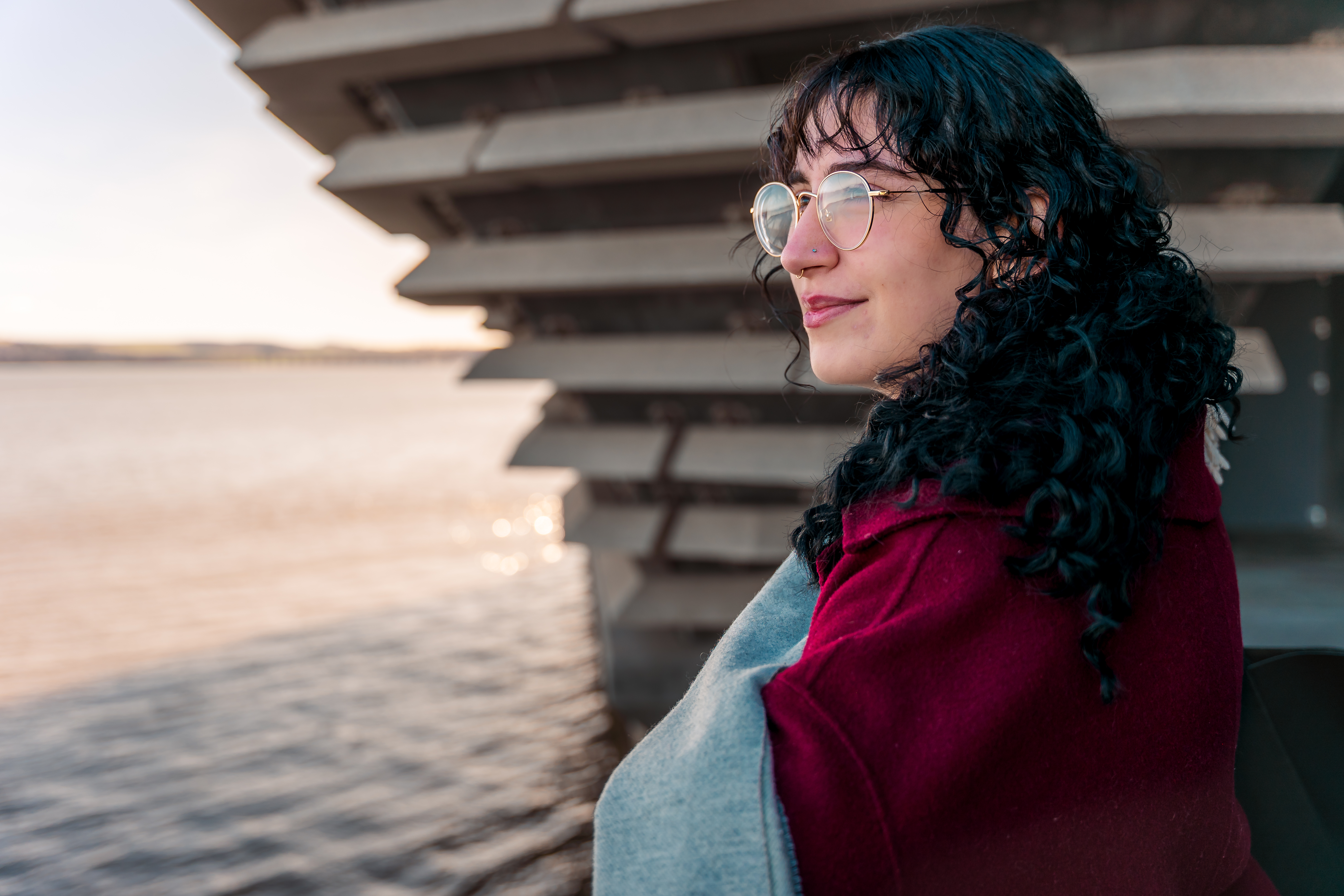 A woman with dark hair and glasses wearing a red coat and grey scarf, she looks off to the distance on the left, in front of her is the Dundee waterfront.