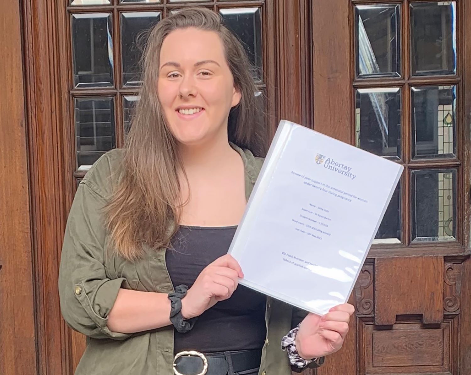 Photo of Libby Todd, a smiling woman with medium‑length brown hair, against a a brown wooden and glass door. She holds her dissertation paper. 